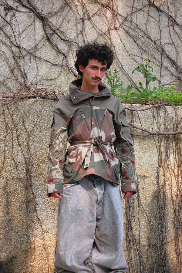 Front view of a camouflage field jacket rebuilt from mismatched panels, cinched with a fabric sash over pale jeans.